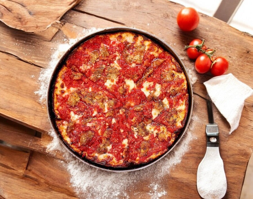 A Labriola Chicago Italian Sausage Deep Dish Pizza, 12-inch and featuring buttery crust with mozzarella cheese and tomato sauce, sits in a round pan on a wooden table. Fresh tomatoes and a spatula complete the classic scene of this inviting Labriola Chicago pizza.