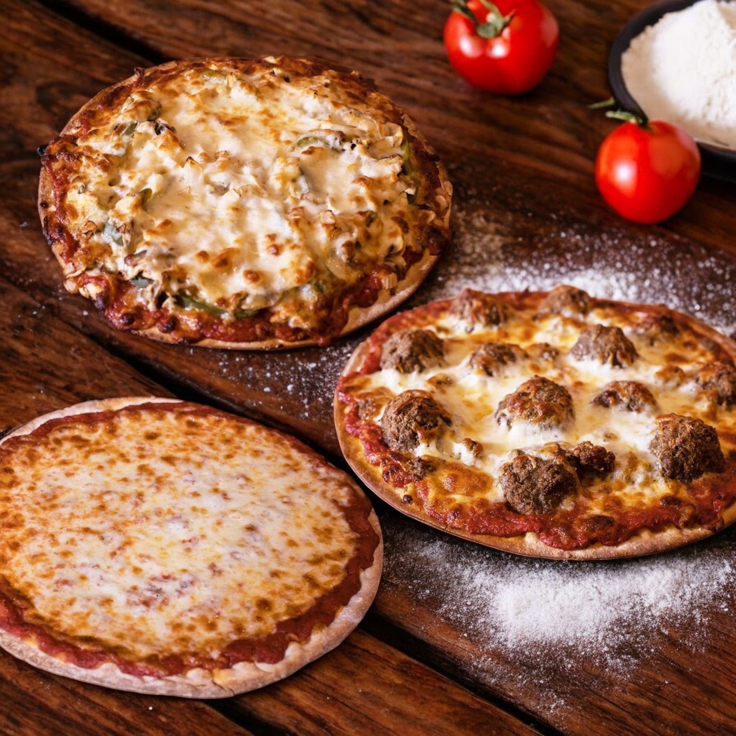 The Bartolini's Pizza 3 Count Combo from Easy Lunches offers a delightful trio on the wooden table: a cheese pizza, one with savory meatballs, and another packed with toppings. Two ripe tomatoes and a flour dish in the background complete this tempting scene of classic pizza flavors.