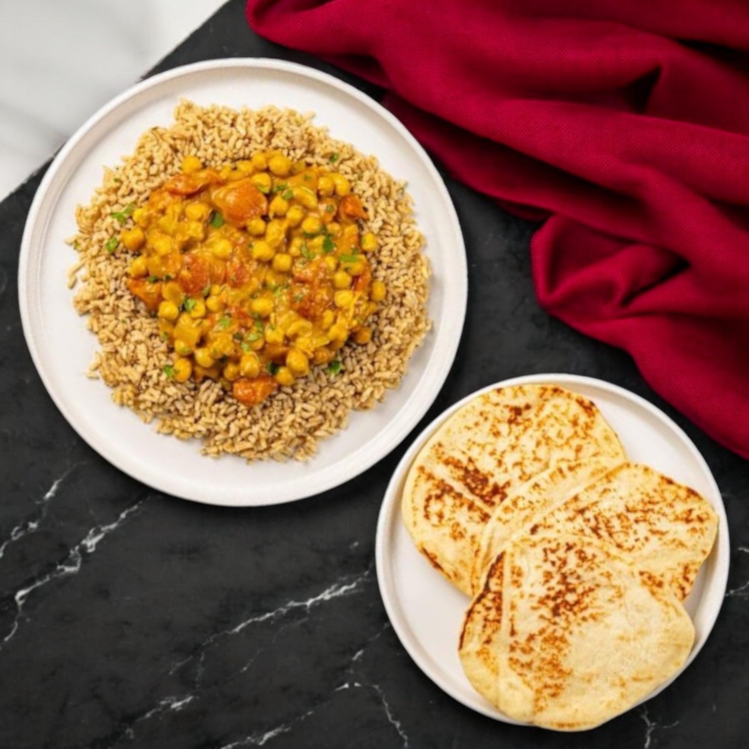 A plate with brown rice topped with FoodHaul's Chana Masala by Jay Farley, featuring chickpeas, tomatoes, and peas. Beside it, a smaller plate holds three slices of toasted flatbread against a red cloth backdrop. This delightful plant-based meal is courtesy of FoodHaul.