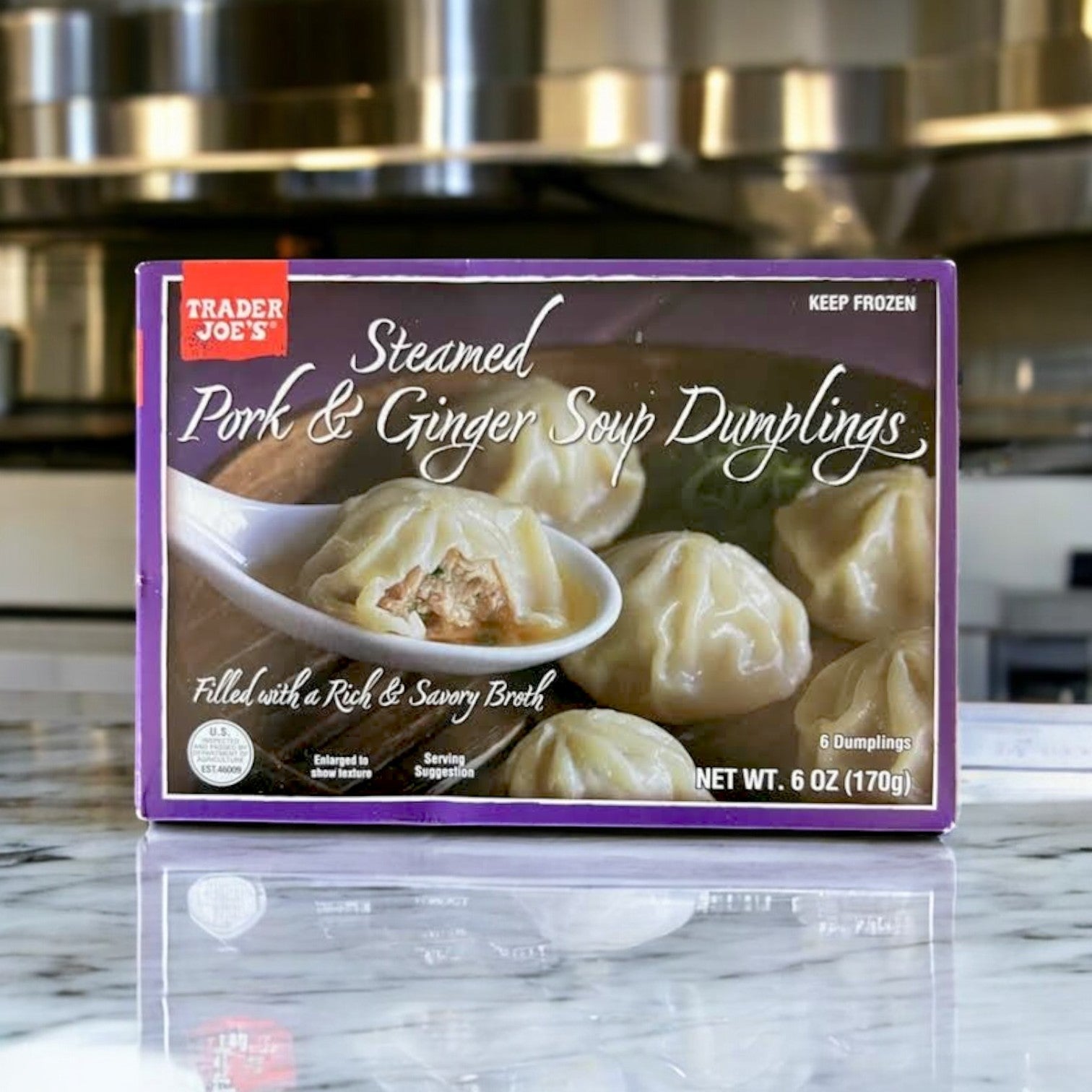 A box of Easy Lunches Steamed Pork & Ginger Soup Dumplings – 6 oz. sits on a marble countertop, displaying its six hand-folded dumplings with traditional Asian flavors on the packaging.