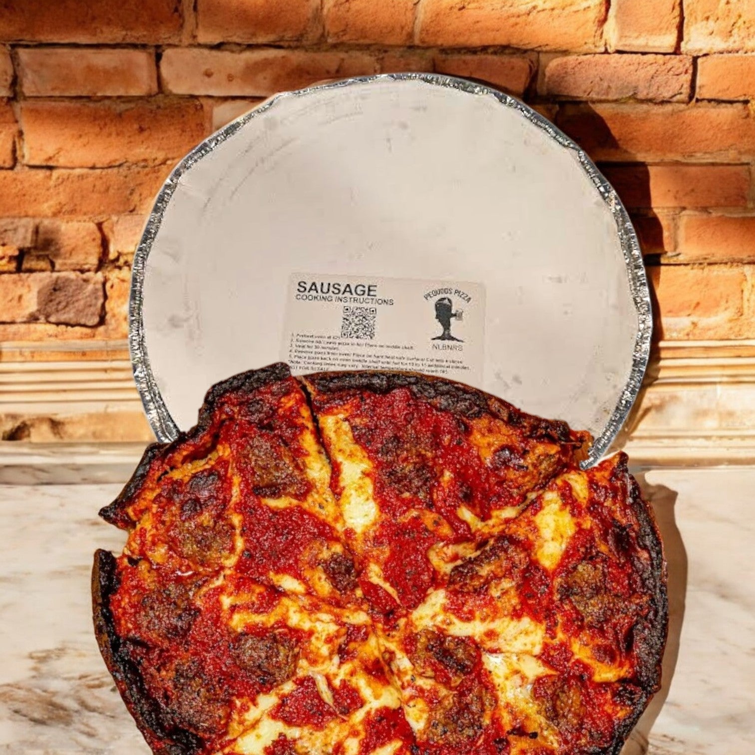 A Pequods Pizza, Sausage Deep Dish 2" tall is set on a marble surface, peeking from its silver foil container in front of a brick wall, reflecting Pequod Pizza’s rich Chicago-style tradition with its charred crust.