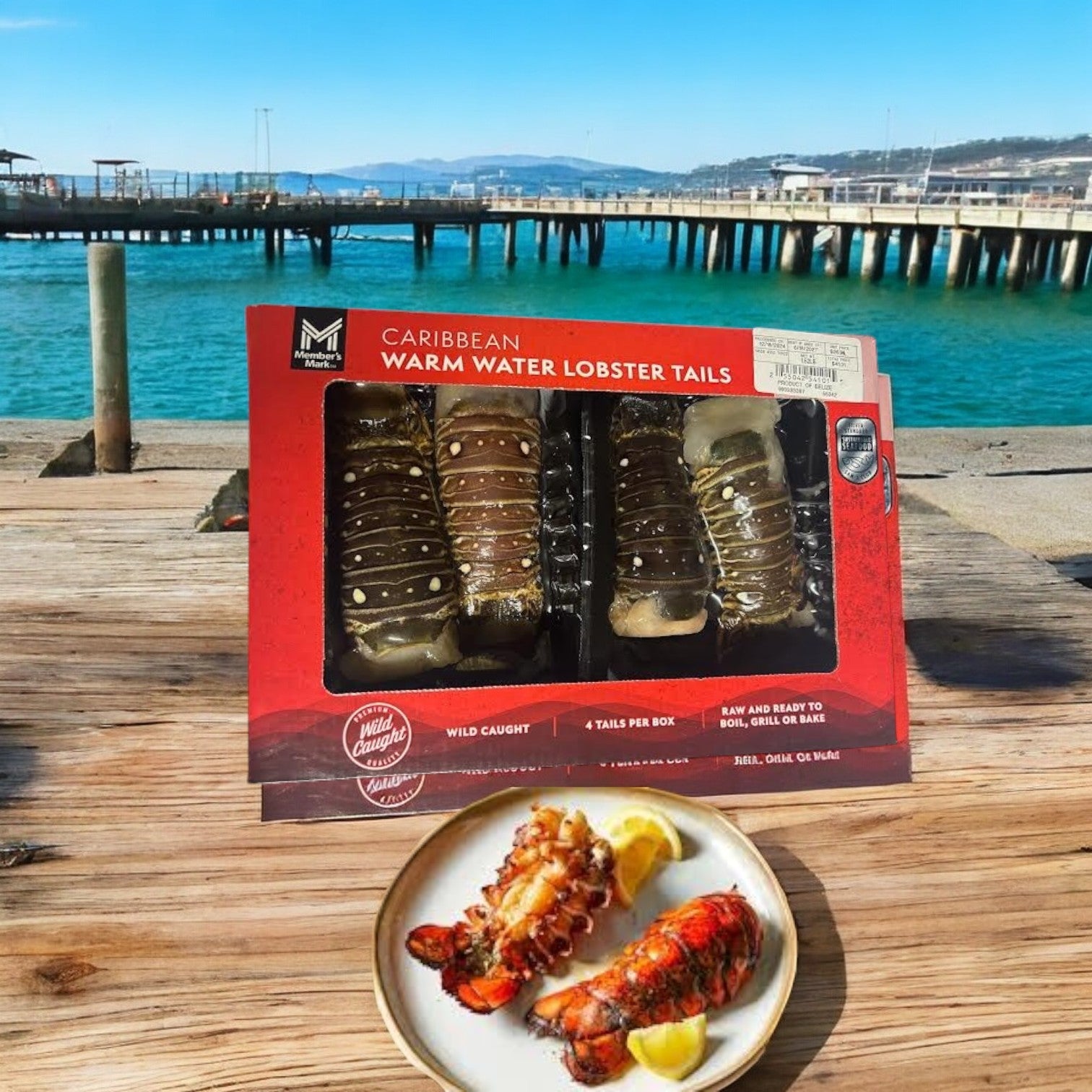 A package of Member's Mark Warm Water Lobster Tails, Frozen, 4 Count-1 Pack, is on a wooden table by the sea, complemented by a small plate of cooked lobster tails and lemon slices.