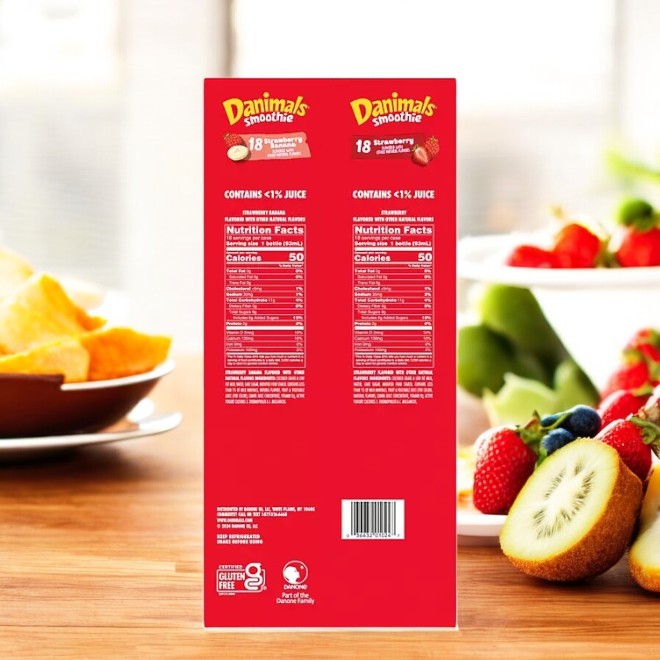 A red box of Dannon Danimals Smoothies Strawberry Variety Pack, complete with a nutrition label, sits beside a fruit bowl of strawberries, blueberries, and kiwi slices on a wooden surface. A perfect nutritious snack for kids.