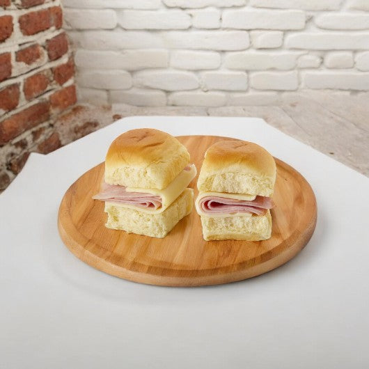 Two small E.A. Sween Smoked Ham and Swiss Sliders are on soft rolls, placed on a round wooden platter against a brick and white backdrop.