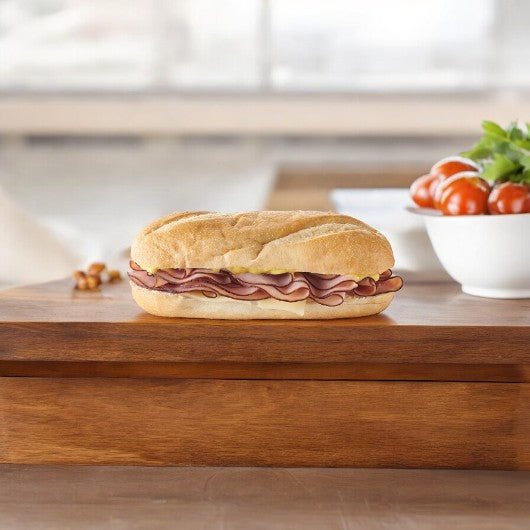 A Smoked Ham & Swiss Cheese Sub from E.A. Sween lies on a wooden surface, with a bowl of tomatoes and lettuce artfully blurred in the background.