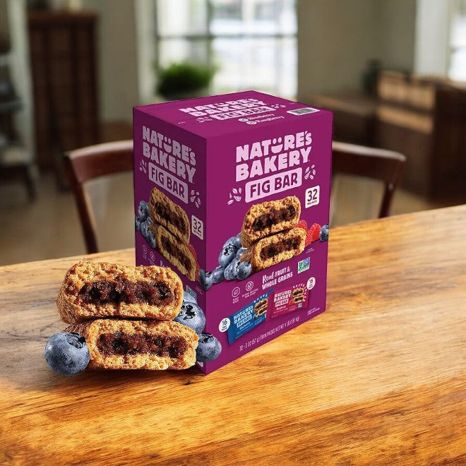 A box of Nature's Bakery Fig Bars, Variety Pack-32 Count, is enticingly displayed on a wooden kitchen table alongside vibrant bar images and fresh blueberries.