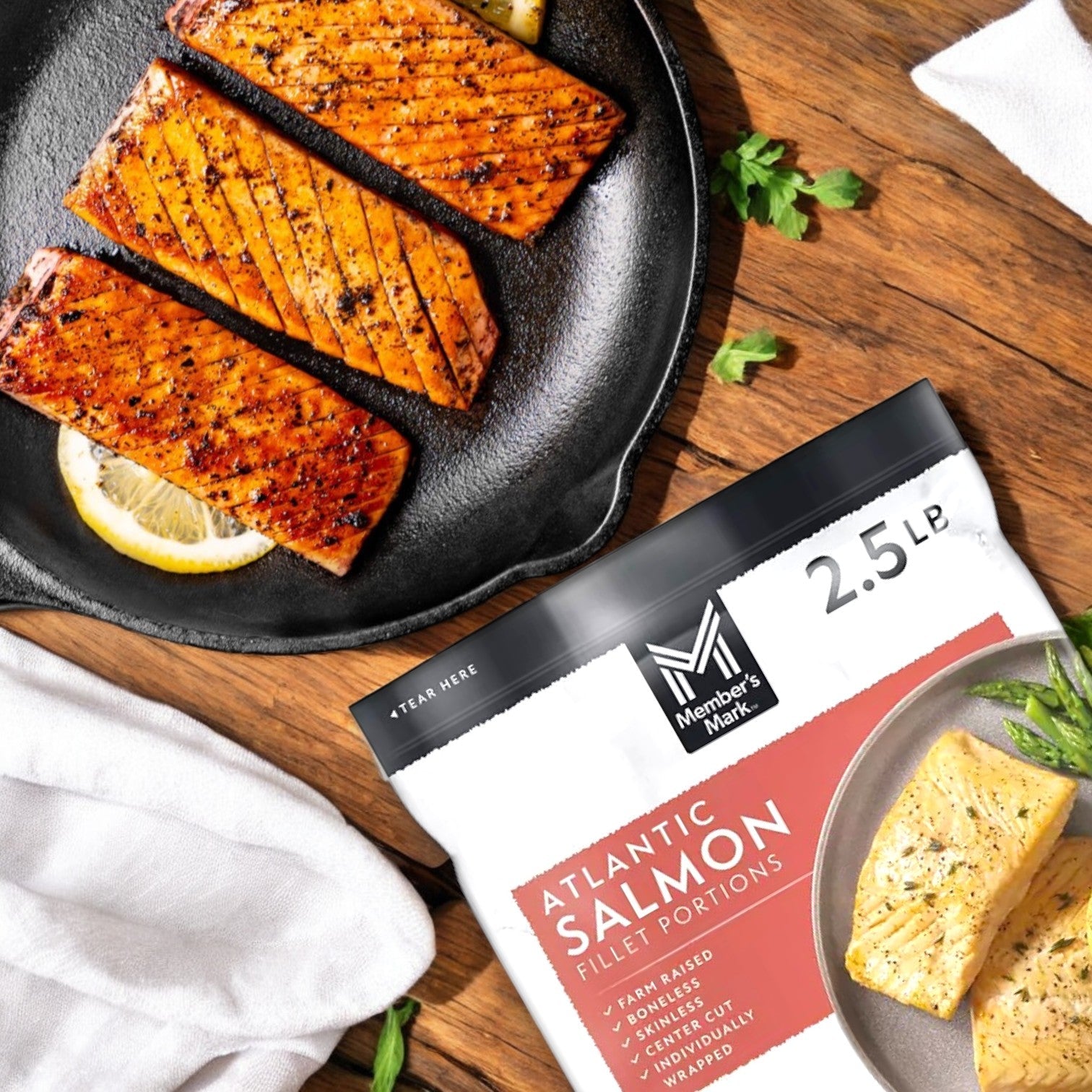 Three sizzling cooked salmon fillets, garnished with lemon slices and herbs, sit beside a Member's Mark Atlantic Salmon Fillet Portions pack (2.5 lb), emphasized by a white cloth, showcasing the omega-3-rich bounty sourced sustainably.