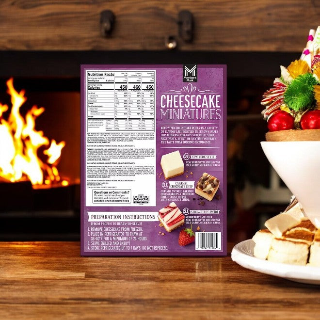 A box of Member's Mark New York Style Cheesecake-3 Count, with instructions, sits by a plate of dessert skewers in front of a cozy fireplace.