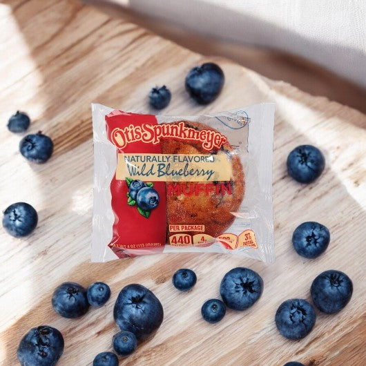 An Otis Spunkmeyer Individually Wrapped Wild Blueberry Muffin, 4 oz., is surrounded by fresh blueberries on a wooden surface.