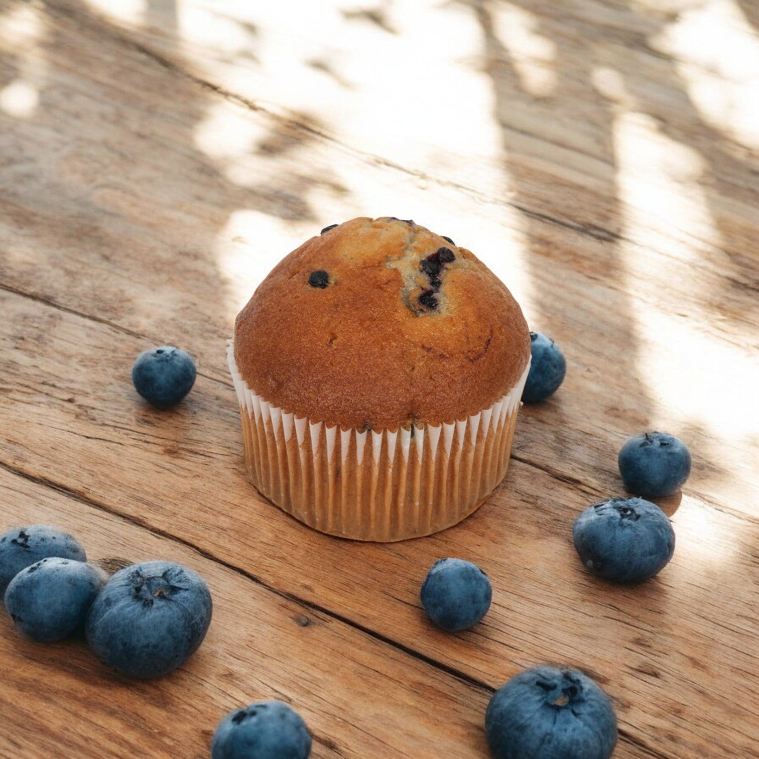 An Otis Spunkmeyer Individually Wrapped Wild Blueberry Muffin, weighing 4 oz., surrounded by scattered blueberries on a rustic wooden surface.
