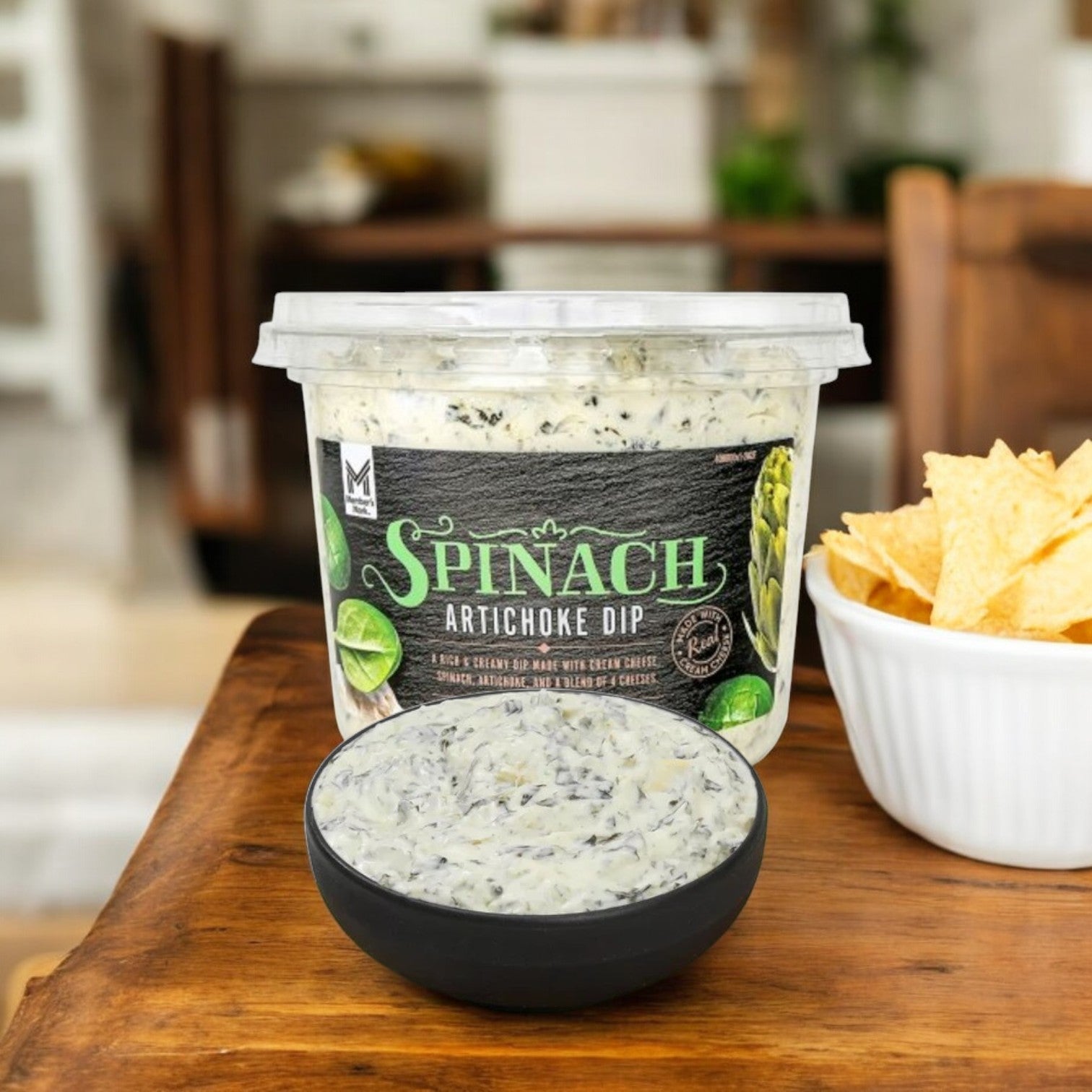 A Member's Mark Spinach Artichoke Dip container (3 count, 6 lbs total) sits invitingly next to a bowl of tortilla chips on a wooden table indoors—a perfect party spread.