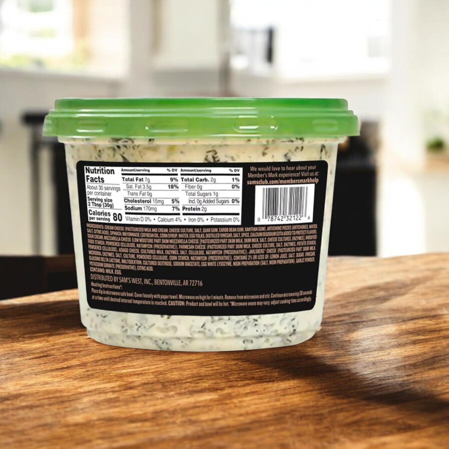 A Member's Mark Spinach Artichoke Dip in a clear plastic container with a green lid rests on wood. The back label shows nutrition facts and ingredients. Ideal for parties, this dip invites indulgence while the background stays elegantly blurred. Comes in 3-pack totaling 6 lbs.