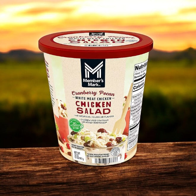A 2 lb container of Member's Mark Cranberry Pecan Chicken Salad sits on a wooden surface with a blurred outdoor background.