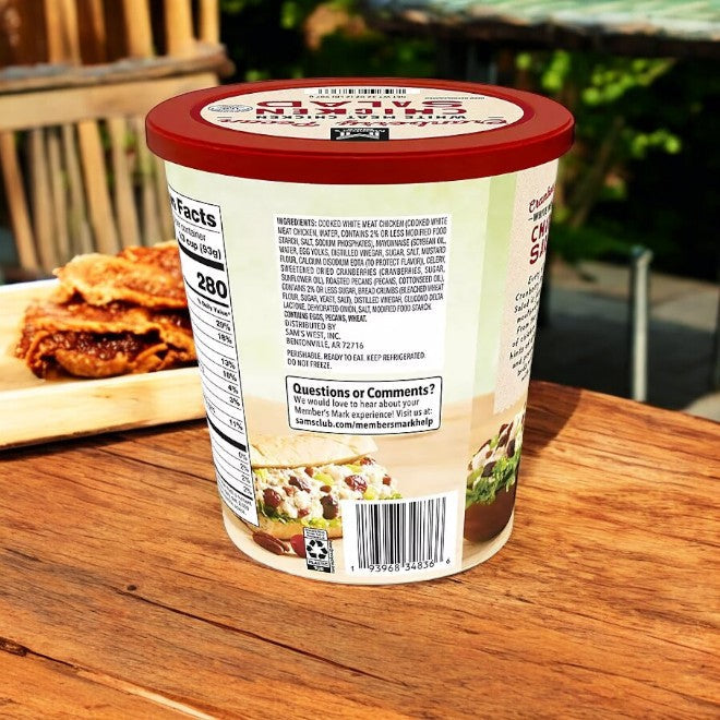 A 2-lb container of Member's Mark Cranberry Pecan Chicken Salad sits outdoors on a wooden table, its red lid and label visible beside a plate, highlighting this tasty ready-to-eat meal from Member's Mark.