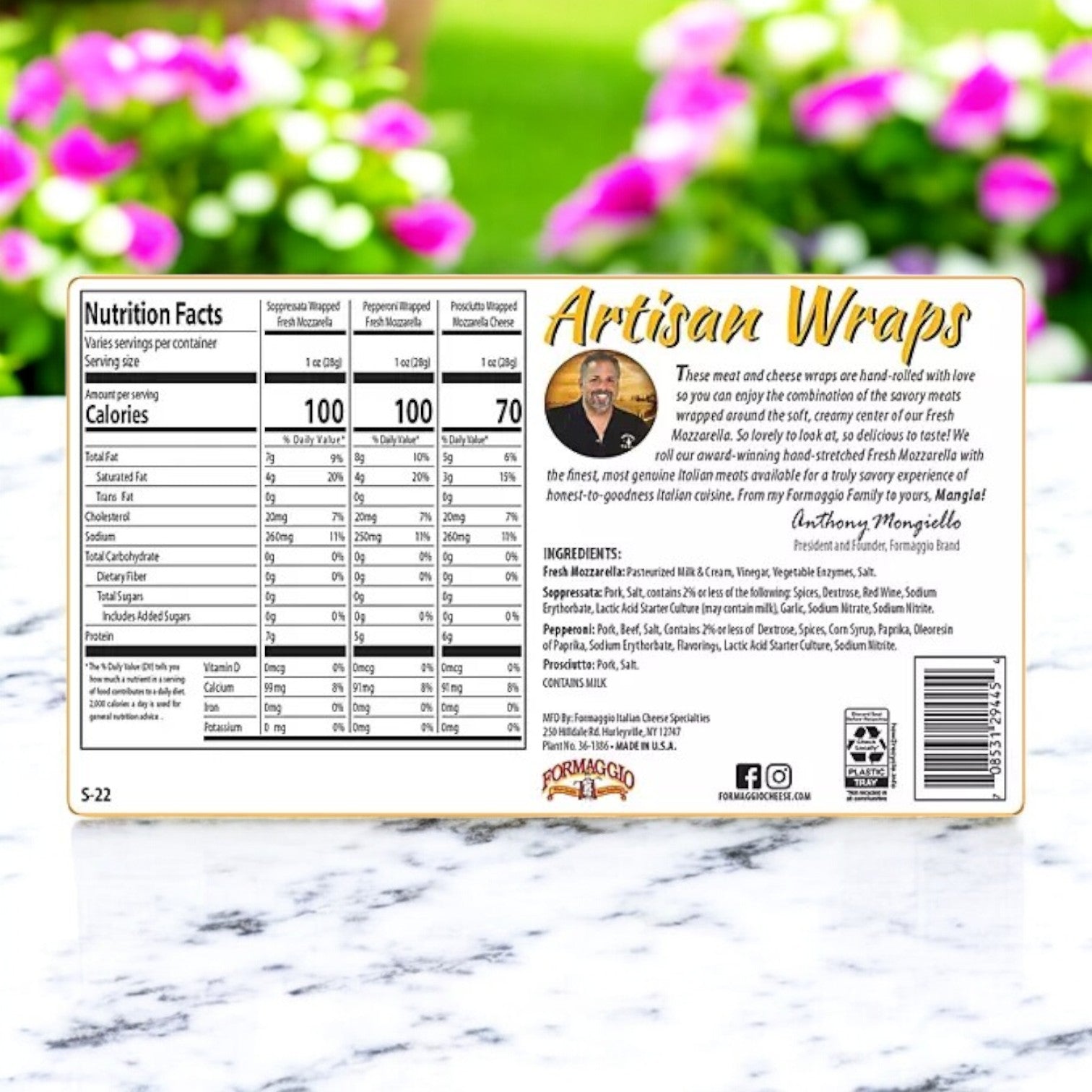 The packaging of Formaggio Cheese Artisan Meat & Mozzarella Variety Wraps, 22 oz, includes nutrition facts, ingredients, and a heartfelt family heritage note. The background features blurred greenery and pink flowers.