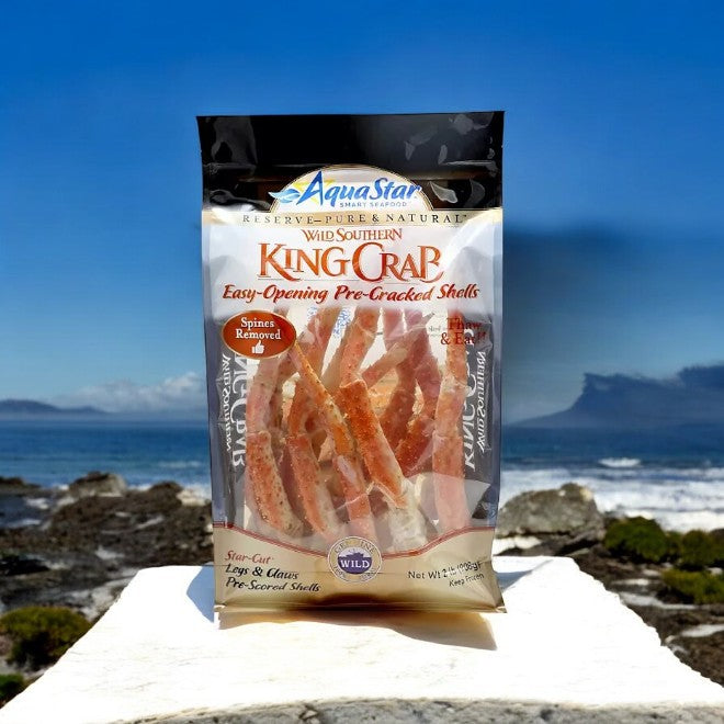 A 2-pound package of Aqua Star Wild Southern King Crab Legs and Claws, frozen, is displayed outdoors on a rocky beach with the ocean in the background, highlighting sustainably sourced premium seafood.