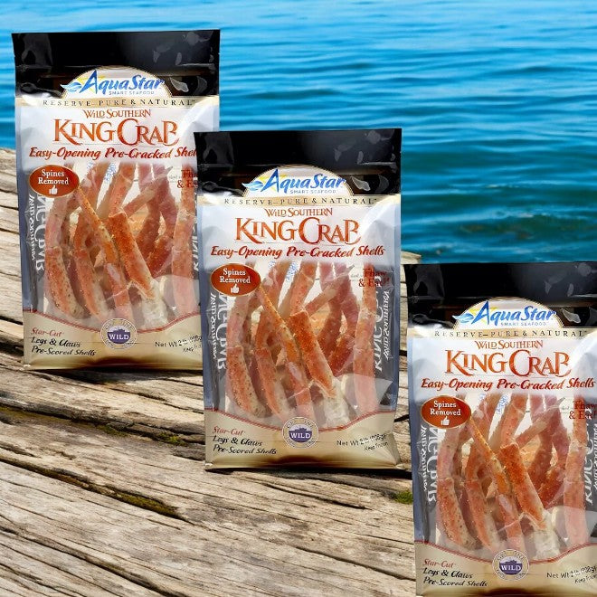 Three cases (6 lbs total) of AquaStar Wild Southern King Crab Legs and Claws, sustainably sourced and pre-cracked, are displayed on a wooden surface by the water.