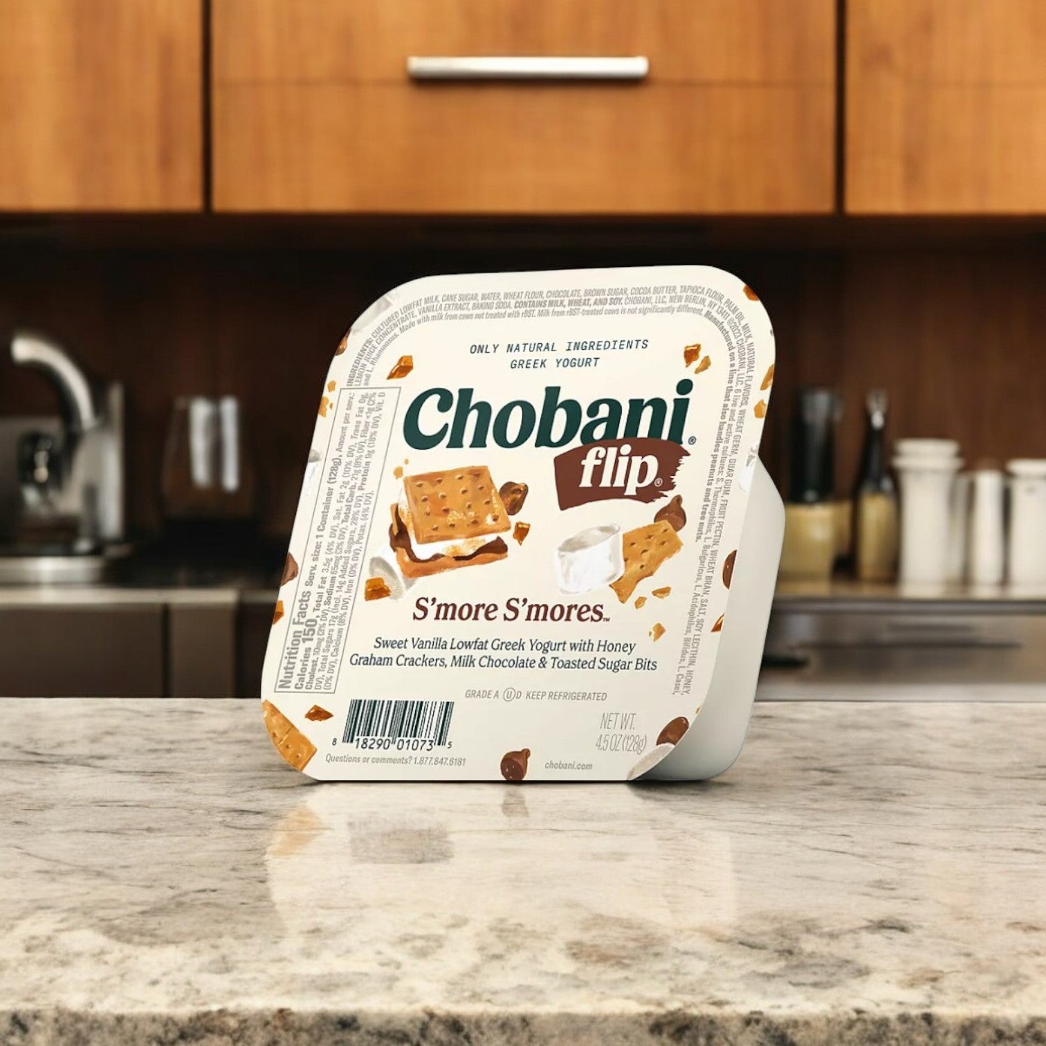 A Chobani Flip Lowfat Greek Yogurt container from a Variety Pack rests on a kitchen counter, set against wooden cabinets with a silver kettle in the background.
