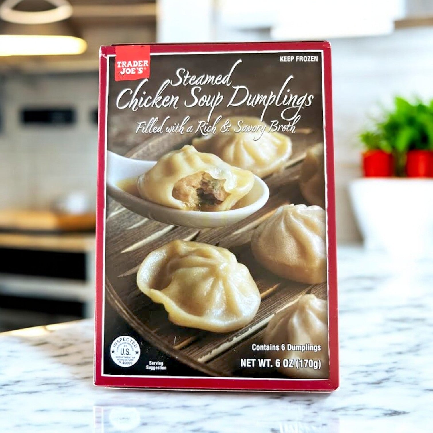 A box of Easy Lunches' Steamed Chicken Soup Dumplings, 6 oz., sits on a marble counter. The packaging displays dumplings on a spoon and describes them as having a rich, savory broth ideal for pairing with versatile ingredients like green pepper julienne to enhance freshness.