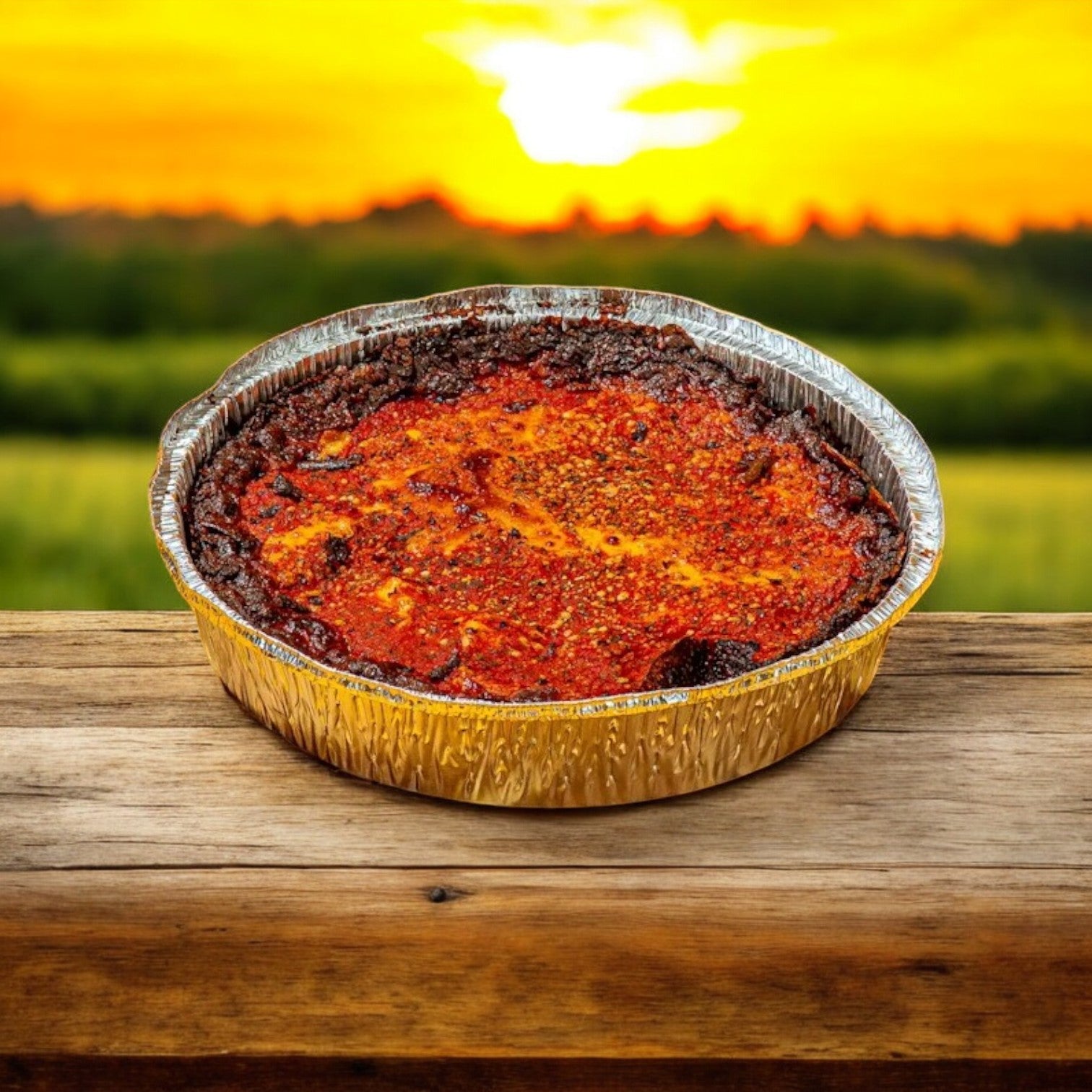 A Pequods Pizza Cheese Deep Dish 2" tall, from Pequod Pizza, sits in a round foil tray on wood, with its caramelized crust glowing warmly against a sunset and grassy landscape.