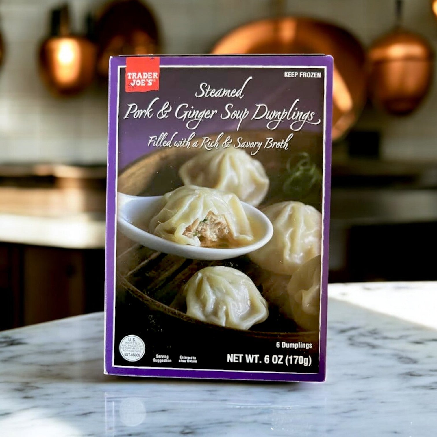 A box of Easy Lunches Steamed Pork & Ginger Soup Dumplings (6 oz.), featuring traditional Asian flavors, sits on a kitchen counter. Copper pans gleam warmly in the background, adding rustic charm to this savory delight.