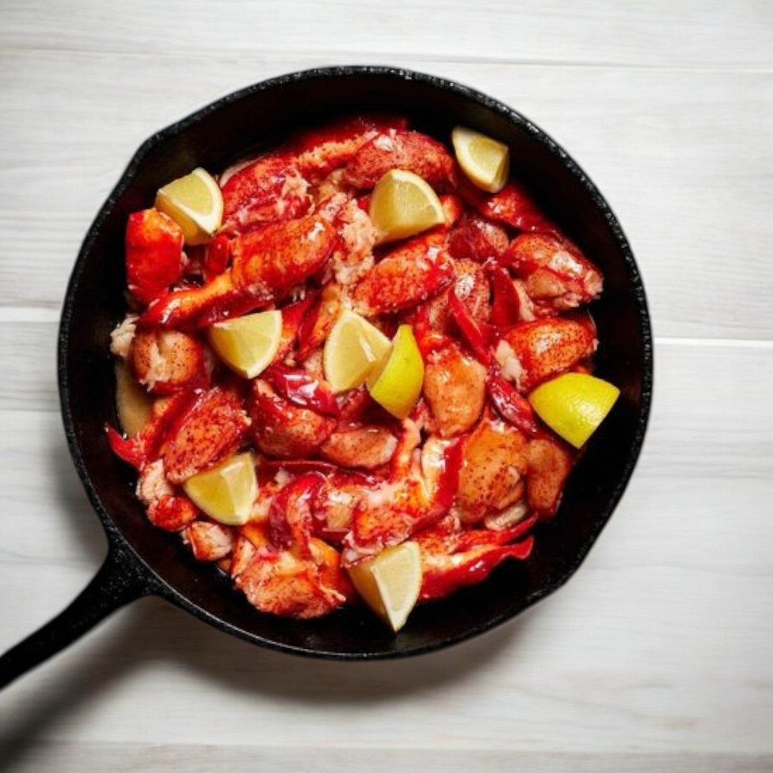 A white wooden surface showcases a skillet filled with Easy Lunches' Maine Lobster Meat Claw and Knuckle-4 Pounds, complete with vibrant lemon wedges.