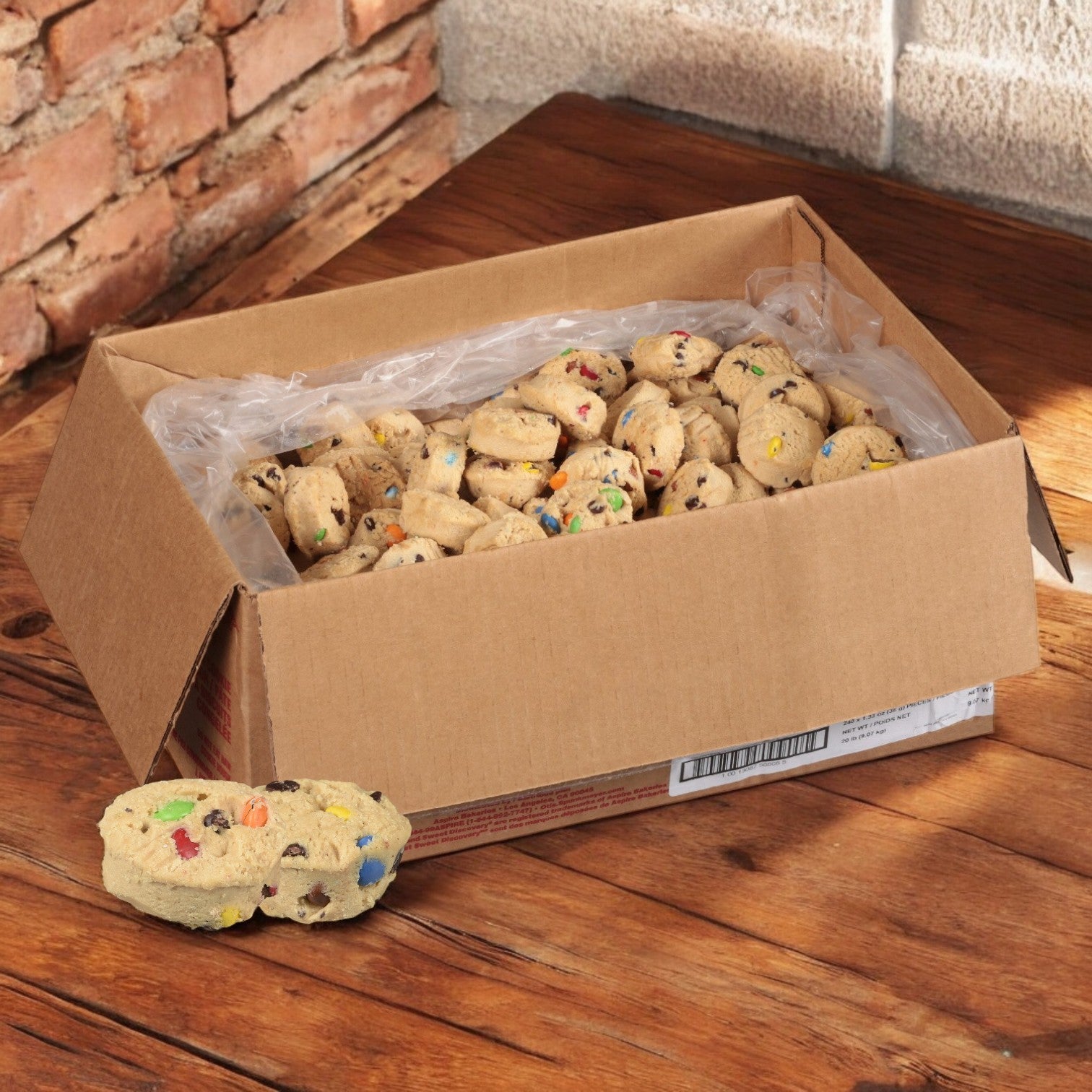 An open cardboard box shows Otis Spunkmeyer Carnival Cookie Dough - 240/1.33 oz, filled with vibrant candy-coated chocolate pieces scattered invitingly on a wooden floor.
