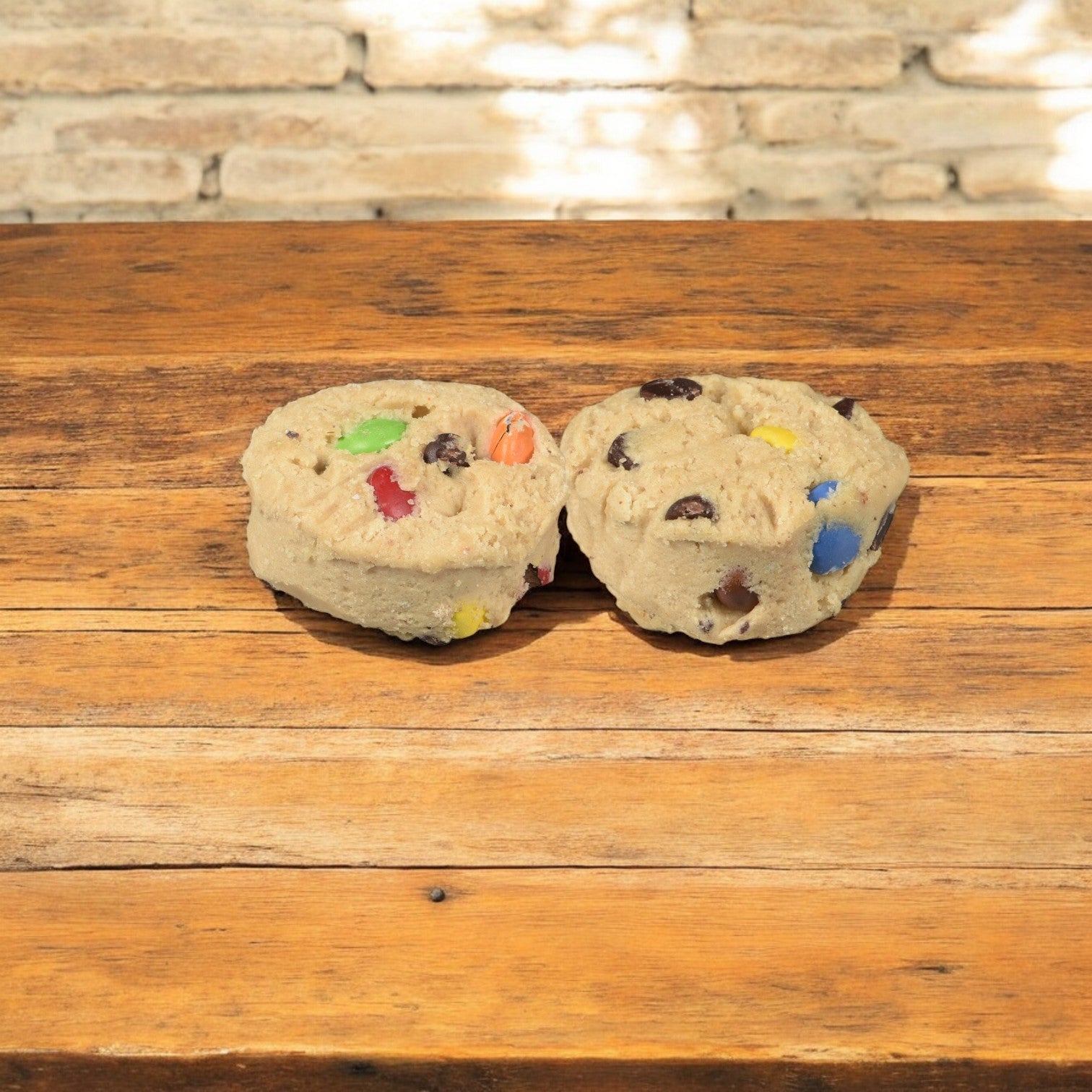 Two Otis Spunkmeyer Carnival Cookie Dough balls, each 1.33 oz and filled with colorful candy-coated chocolate pieces, rest on a wooden table against a brick background.