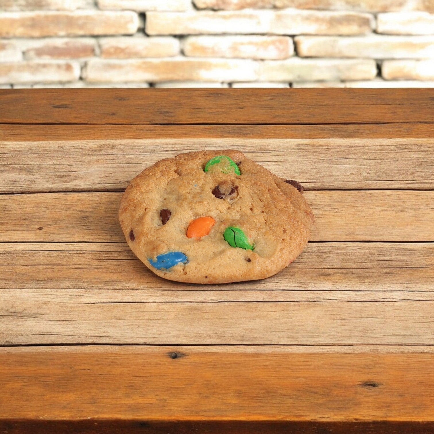 An Otis Spunkmeyer chocolate chip cookie from the Carnival Cookie Dough (240/1.33 oz) rests on a wooden surface against a brick wall, exuding playful vibes.