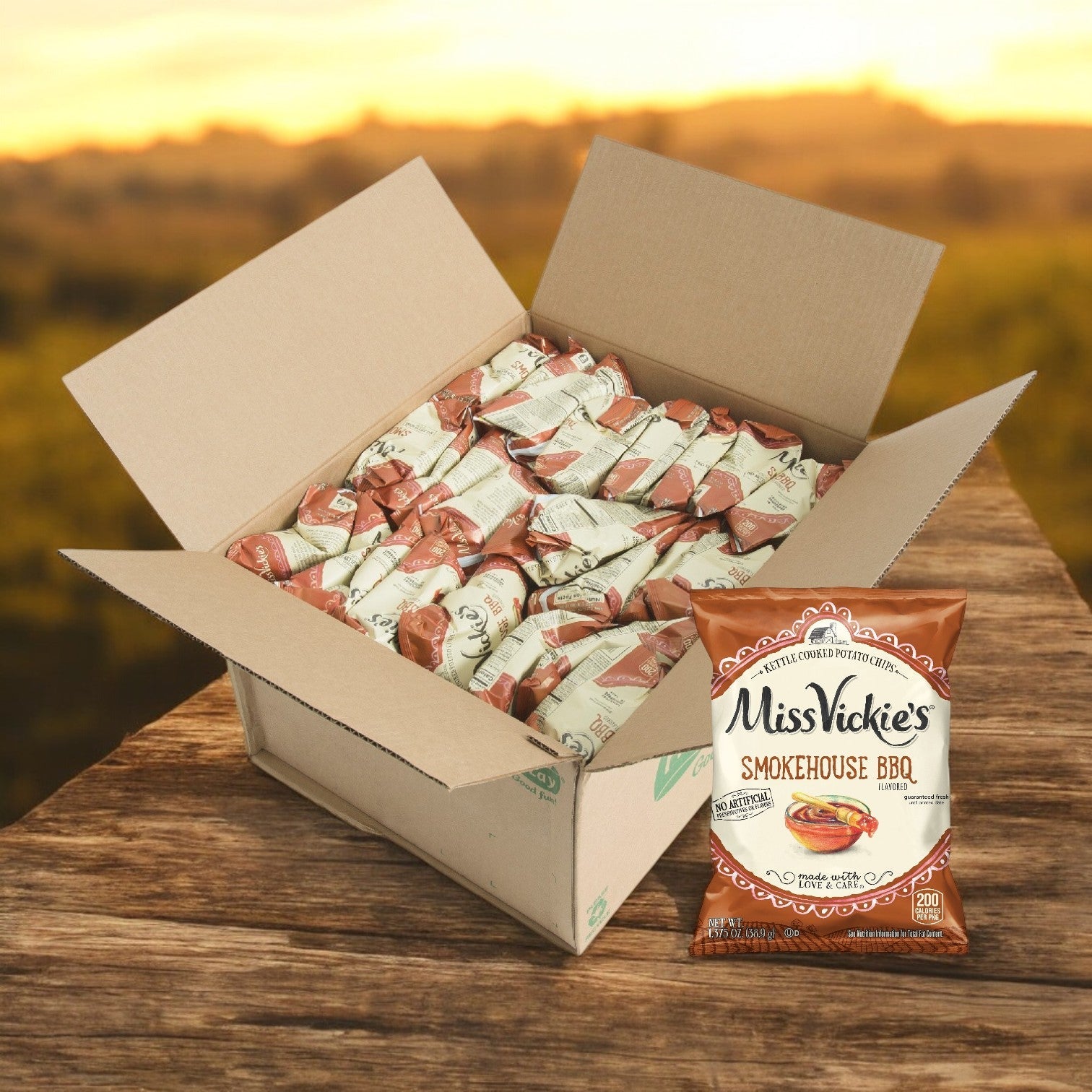 An open box of Miss Vickie’s Potato Chips Smokehouse BBQ Kettle 64/1.375 oz. sits on a wooden surface, with one bag in front, offering a crispy snack experience made from high-quality potatoes.