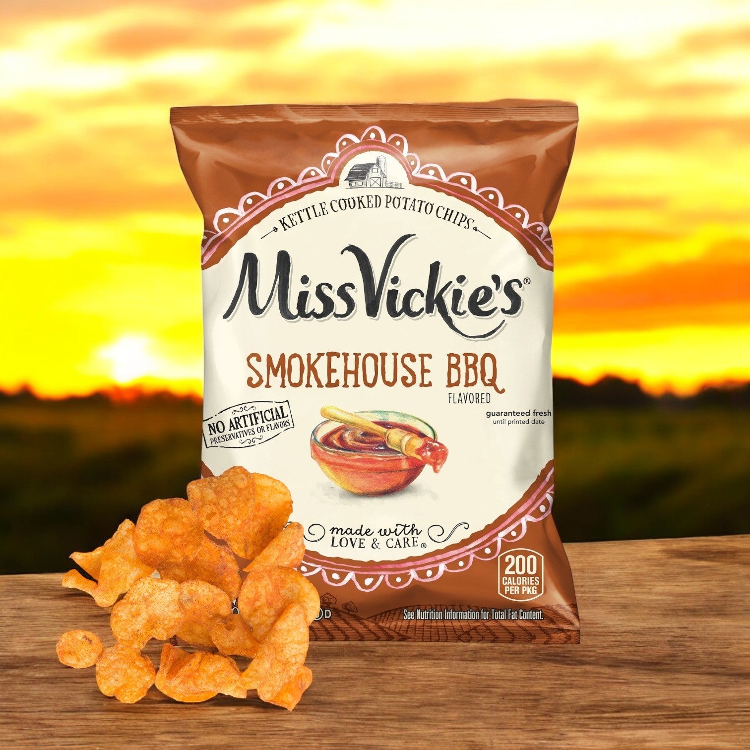 A bag of Miss Vickie’s Potato Chips Smokehouse BBQ Kettle 64/1.375 oz. is shown with scattered chips in front, made from high-quality potatoes, set against a warm sunset over a blurred landscape.