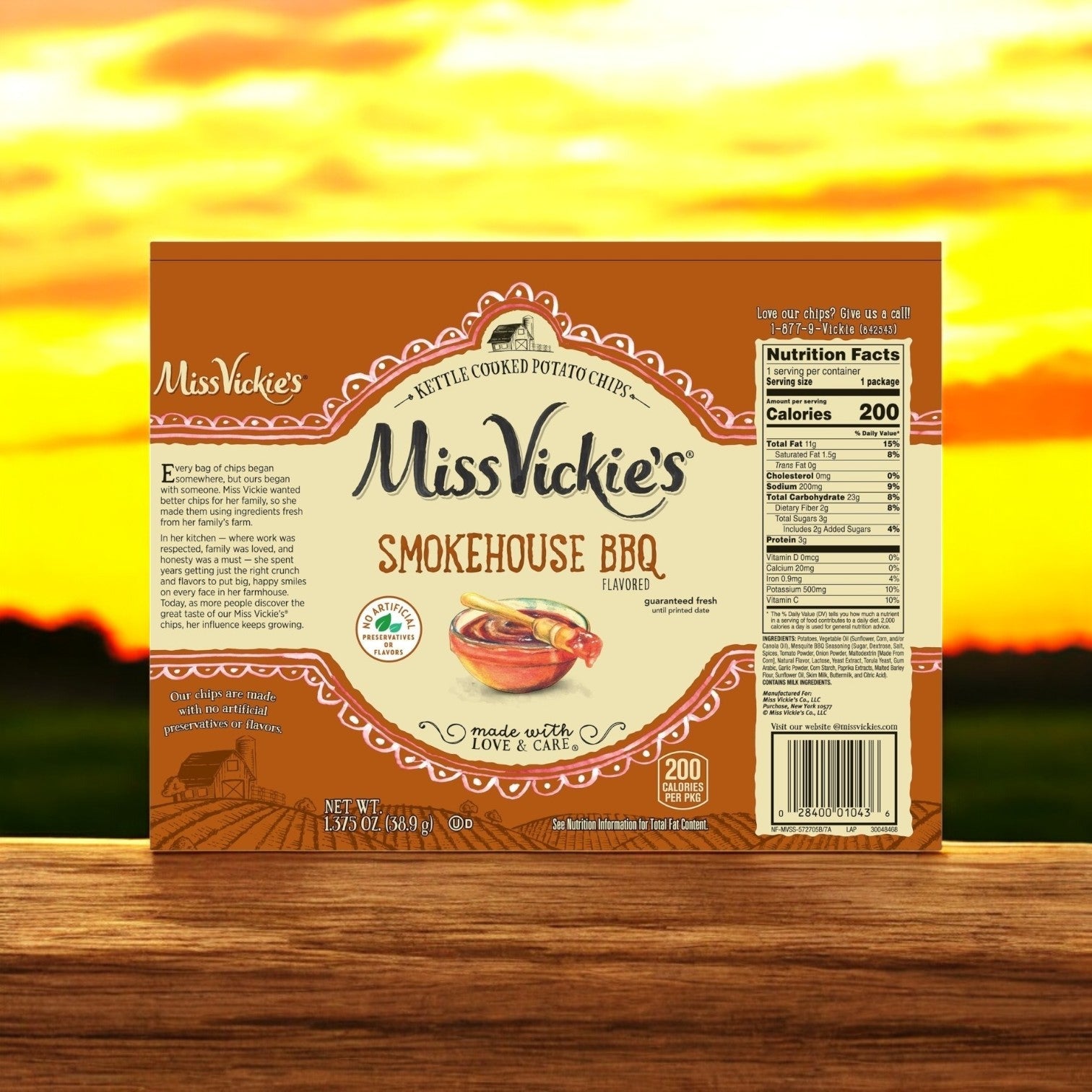 A box of Miss Vickie’s Potato Chips Smokehouse BBQ Kettle 64/1.375 oz., made from premium potatoes, rests on a wooden surface with a sunset in the background.