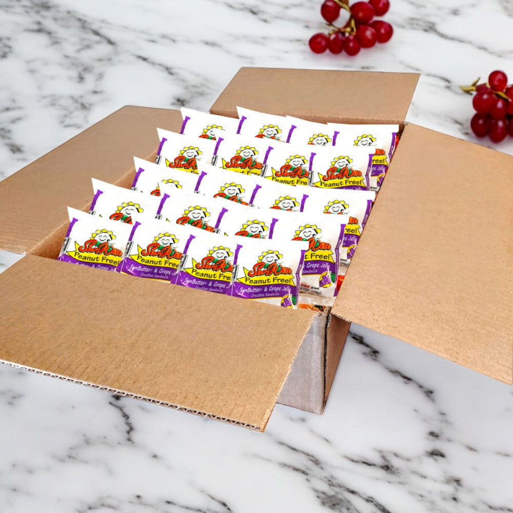 An open cardboard box on a marble surface holds individually wrapped Sunwise Grape Jelly Sandwiches, providing a delightful nut-free snack experience, with 96 sandwiches in the case and red grapes in the background. Enjoy free shipping!.