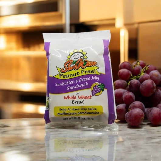 A Sunwise Individually Wrapped Sunbutter & Grape Sandwich, a 5.2-ounce peanut-free option, is on the counter. Nearby are some red grapes.