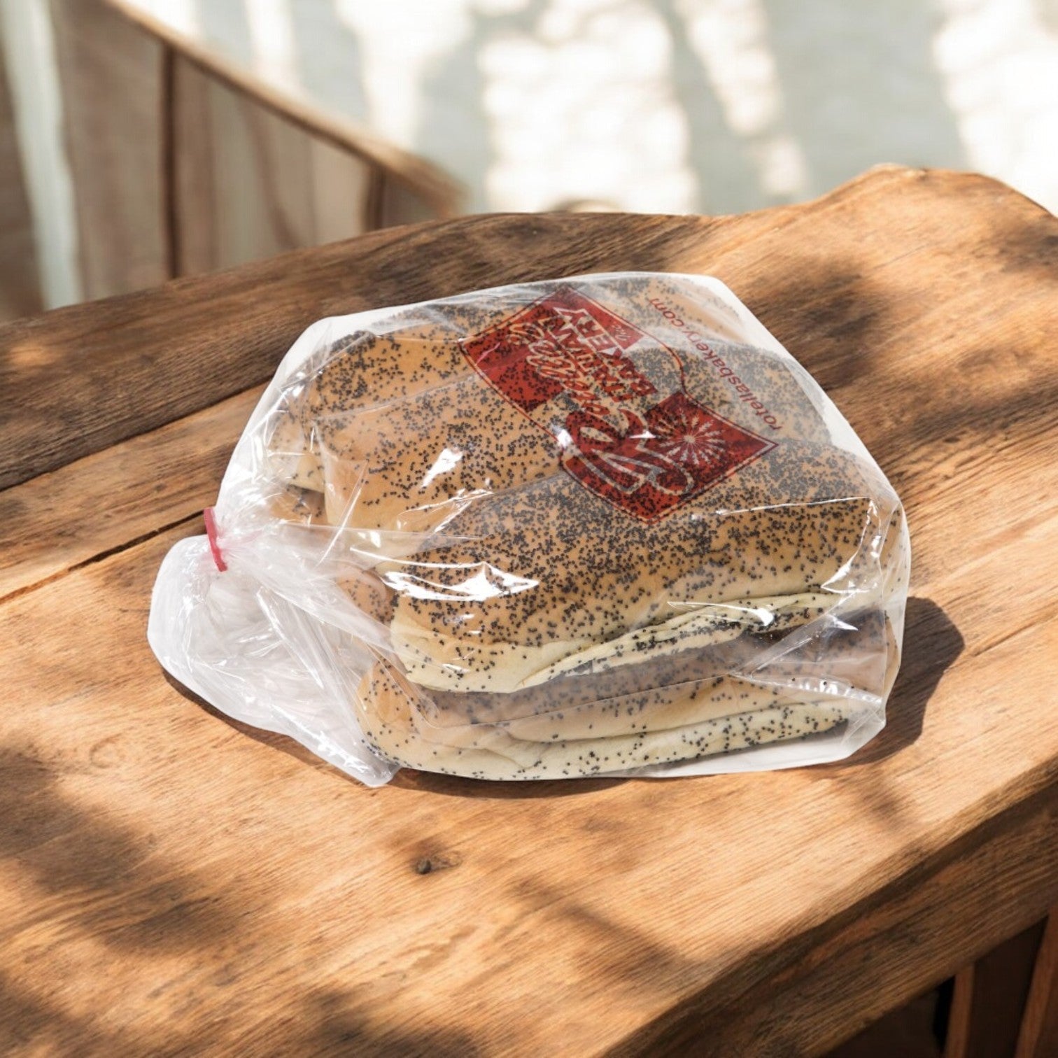 A 6-pack of Easy Lunches' Rotellas Poppyseed Sliced Hot Dog Buns sits in a plastic bag on a wooden table, sunlight creating shadows around it.