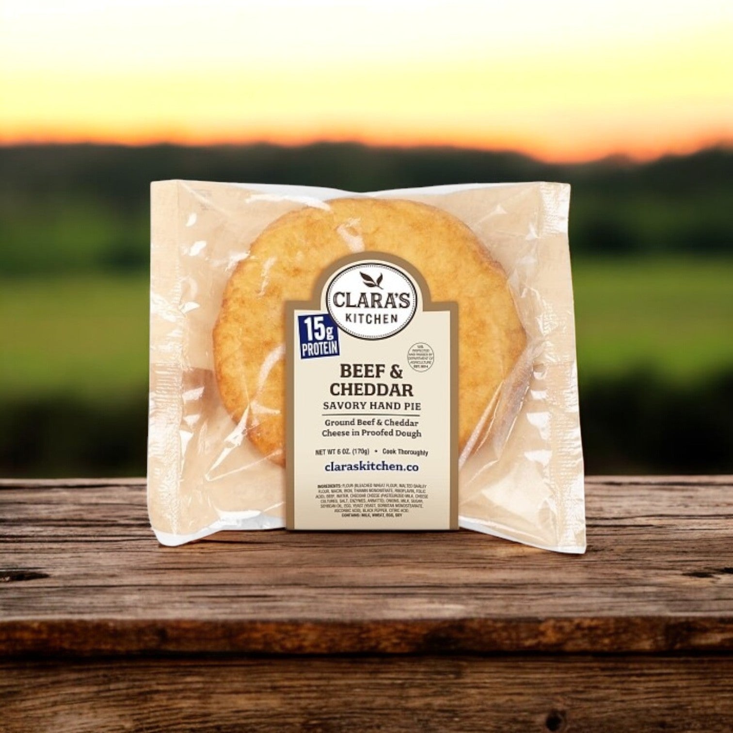 The 6 oz. Clara's Kitchen Beef & Cheddar Handpie with a crispy crust rests on a wooden surface, while blurred outdoor scenery is in the background.