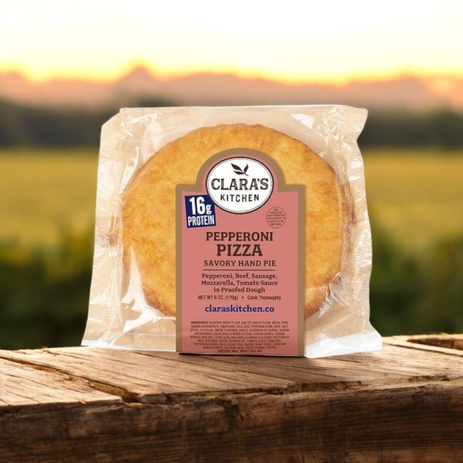 Clara's Kitchen Pepperoni Pizza Handpie, 6 oz., from Clara's Kitchen, is individually packaged and placed on a wooden surface with a blurred outdoor background.