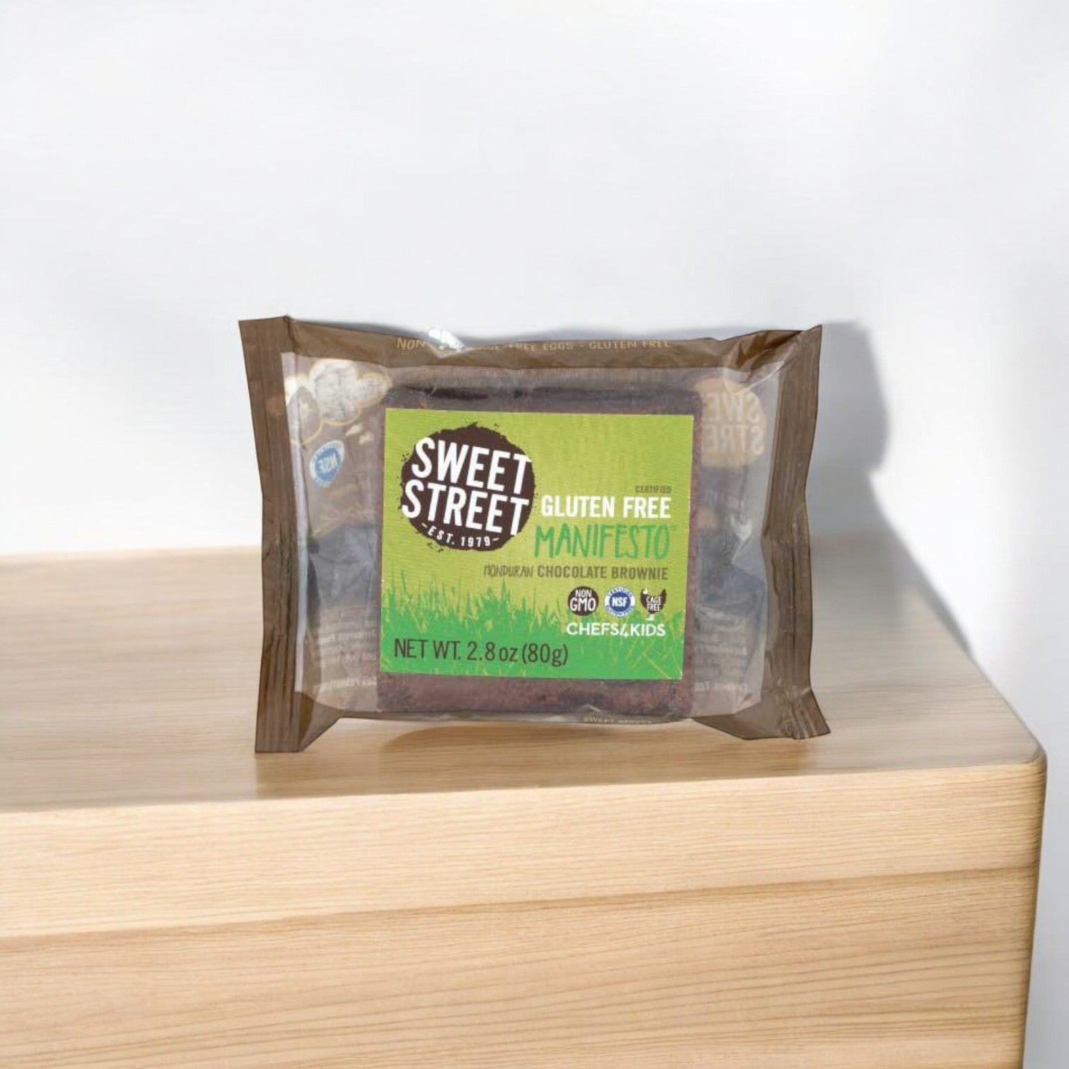 A 2.8 oz. Sweet Street Honduran Chocolate Manifesto Brownie from Sweet Street, showcased with its rich chocolate on a wooden surface, is an ideal addition to any dessert menu. Sold in cases of 48, these fudgy brownies promise a decadent treat without gluten.