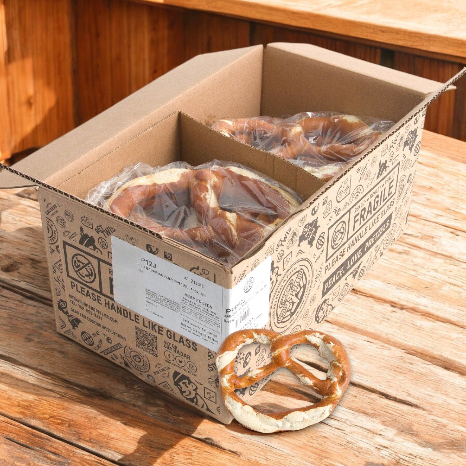 A cardboard box with illustrations holds two Prop & Peller Pretzel Soft Jumbo Pretzels in plastic bags on a wooden surface; one pretzel is outside the box. The packaging is labeled "Fragile: Please handle like glass.