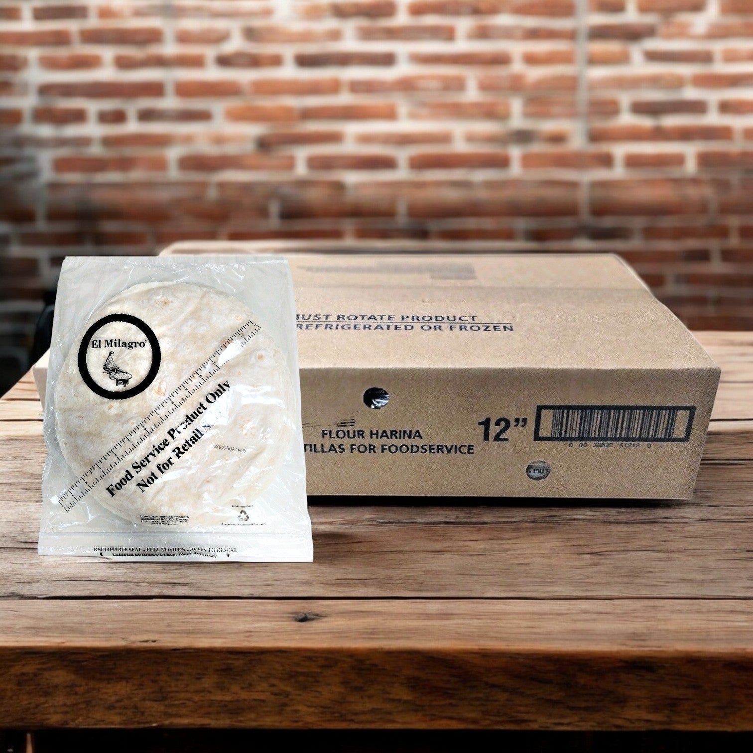 In front of a large foodservice-labeled box, a pack of El Milagro Flour Tortillas - 12-Inch rests on a wooden table, set against a backdrop of rustic brick walls, emphasizing their authentic quality.