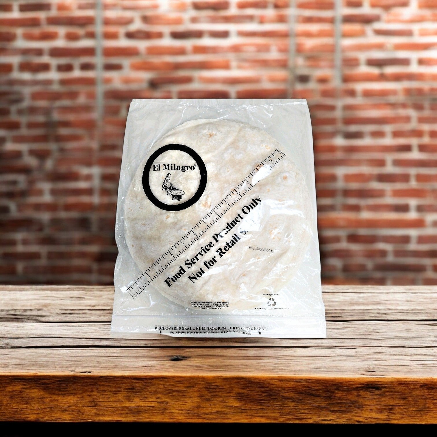A clear plastic package of El Milagro Flour Tortillas - 12-Inch - 12 Packs of 12 Dozen lies on a wooden surface with a rustic brick wall backdrop, highlighting their authentic quality.