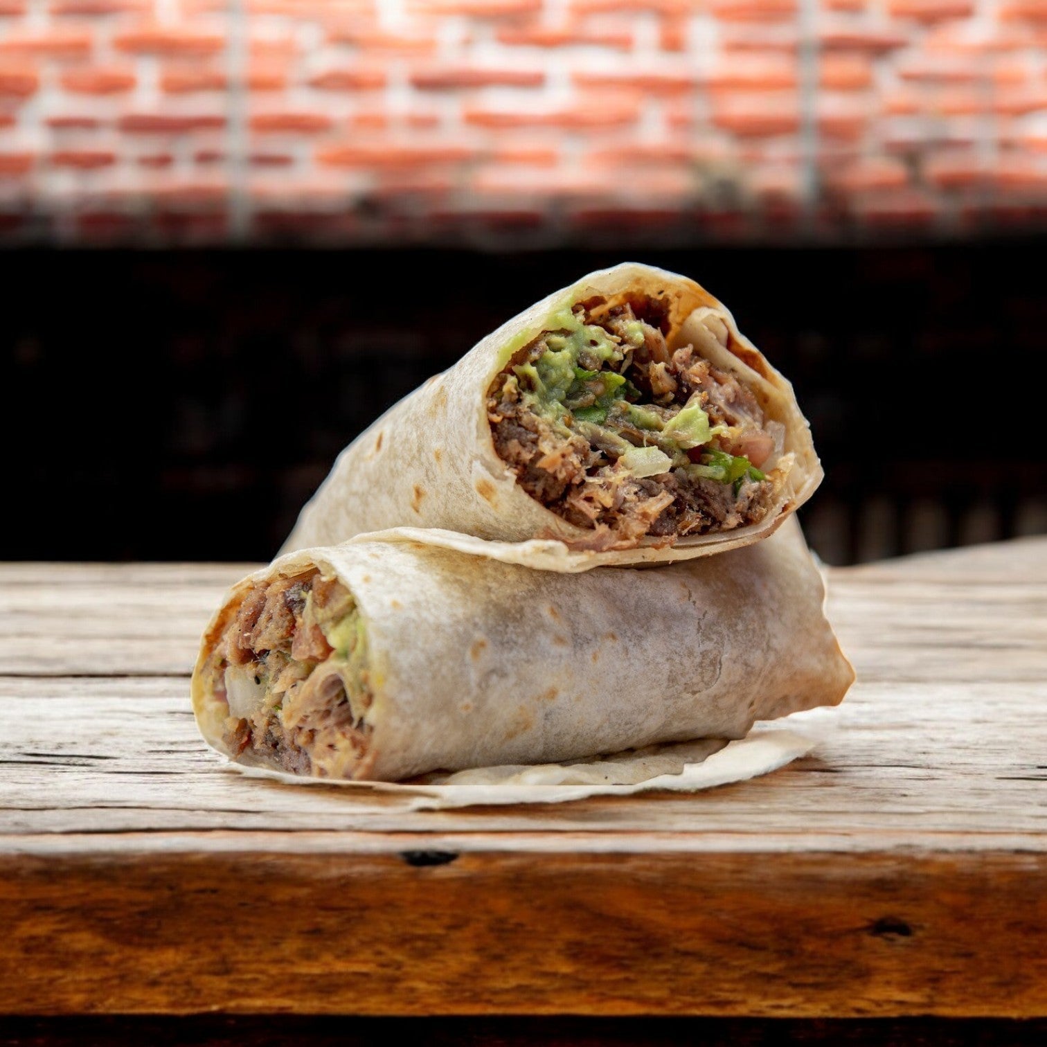 Two halves of a burrito with meat and guacamole are stacked on a wooden table against a brick wall, highlighting the authenticity of El Milagro Flour Tortillas - 12-Inch, available in 12 packs.