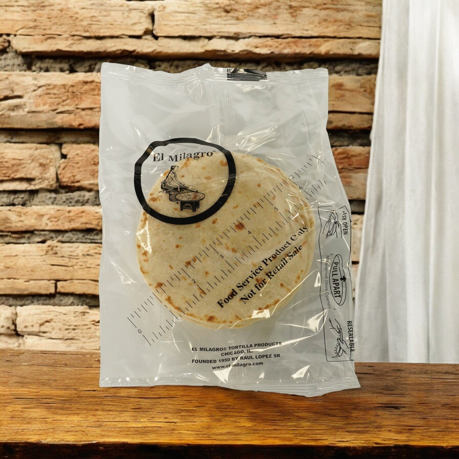 A sealed package of El Milagro White Flour Tortillas (5-inch, 42 packs of 1 dozen) sits on a wooden surface with a brick wall backdrop. Ships Mondays—plan ahead!.