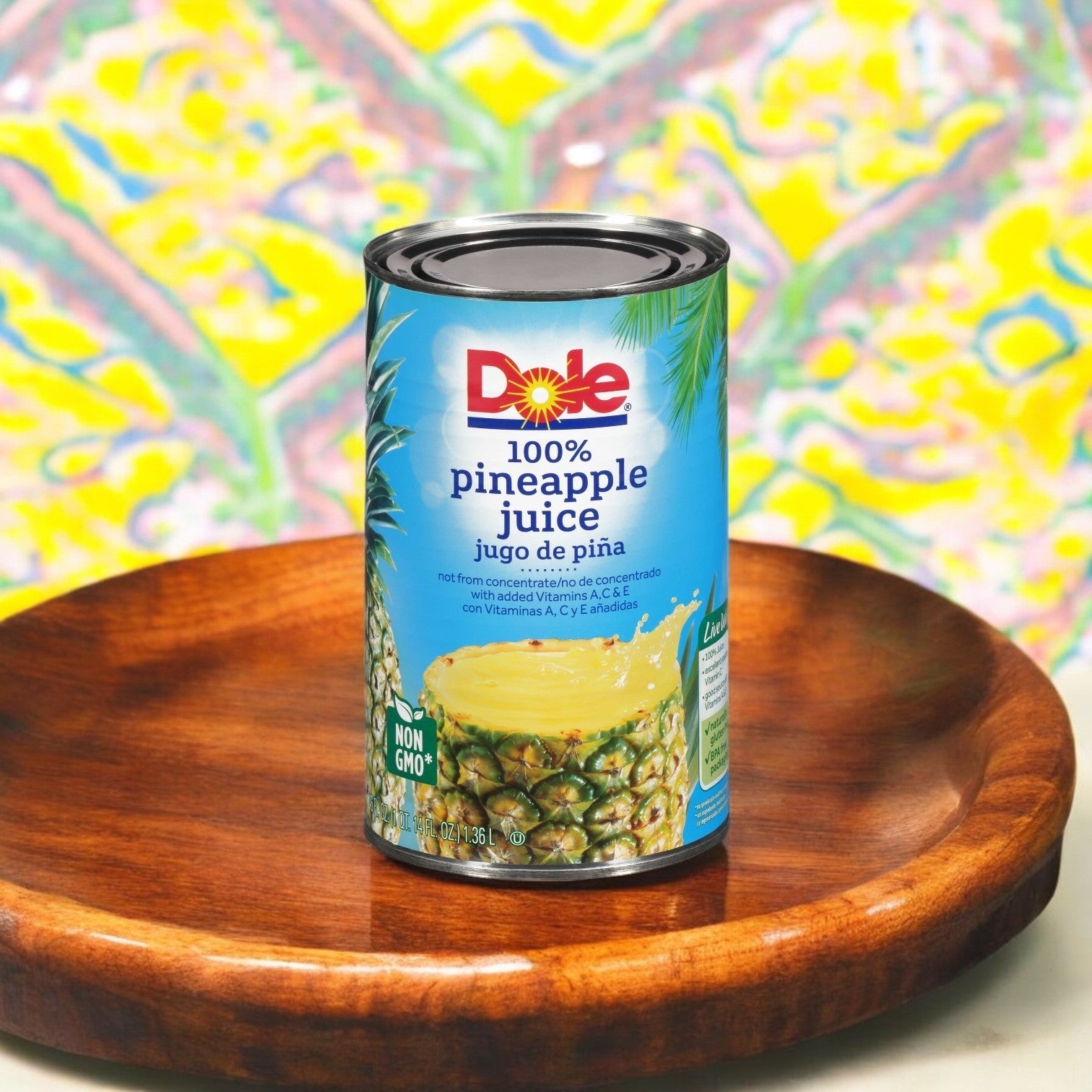 A can of Dole Pineapple Juice, 46 oz. from the 1 Case, 12 Pack line rests on a wooden plate, exuding tropical flavor against a vibrant abstract backdrop.