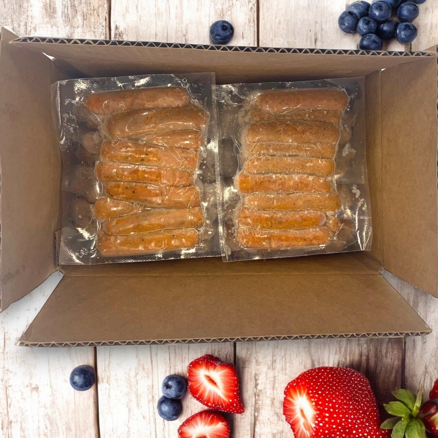 A cardboard box holds ten 1 lb packs of Amylu Chicken Sausage Links, Apple & Maple, made with all-natural ingredients. Vibrant blueberries and strawberries are artfully placed around the box on a wooden surface, enhancing this wholesome scene.