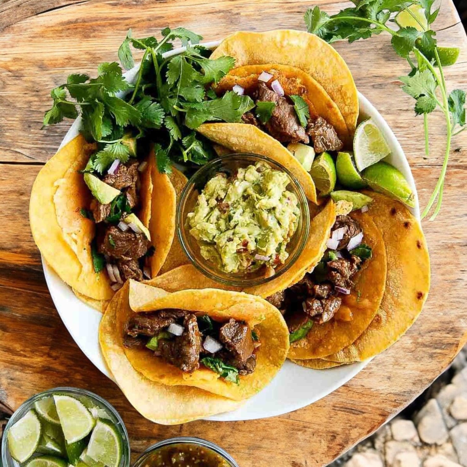 Steak tacos made with El Milagro Yellow Corn Tortillas (6"), topped with onions and avocado, served with guacamole, lime wedges, cilantro, and drinks on a wooden table. Ships Mondays; plan accordingly!.