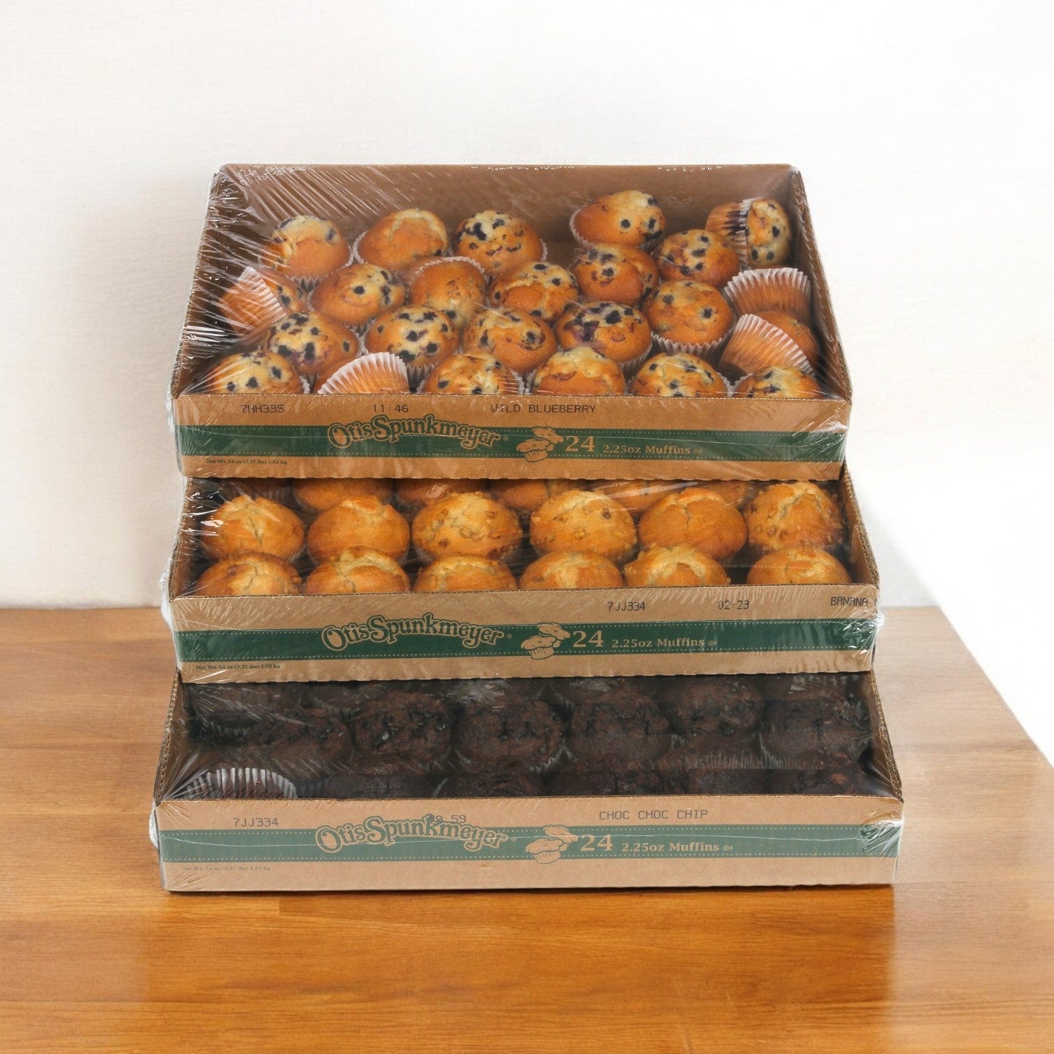 Three trays of Otis Spunkmeyer Muffin Variety 96/2.25 oz., featuring wild blueberry, banana nut, and chocolate chip flavors, stacked on a wooden table. Each Blueberry Banana and Chocolate Chip muffin is individually wrapped in plastic for freshness.