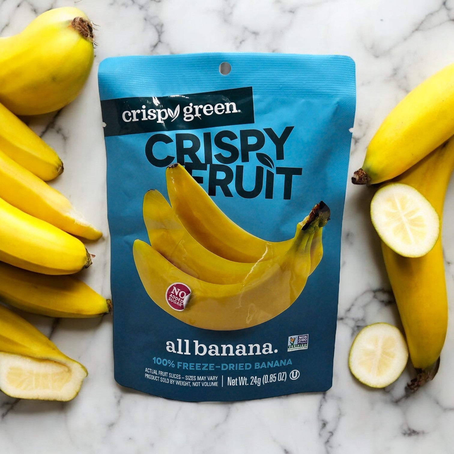 A blue package of Crispy Green Crispy Fruit All Banana sits on marble, surrounded by fresh bananas and slices. Enjoy sustainable snacking with this crunchy treat, ideal with our famous freeze-dried pineapple slices.