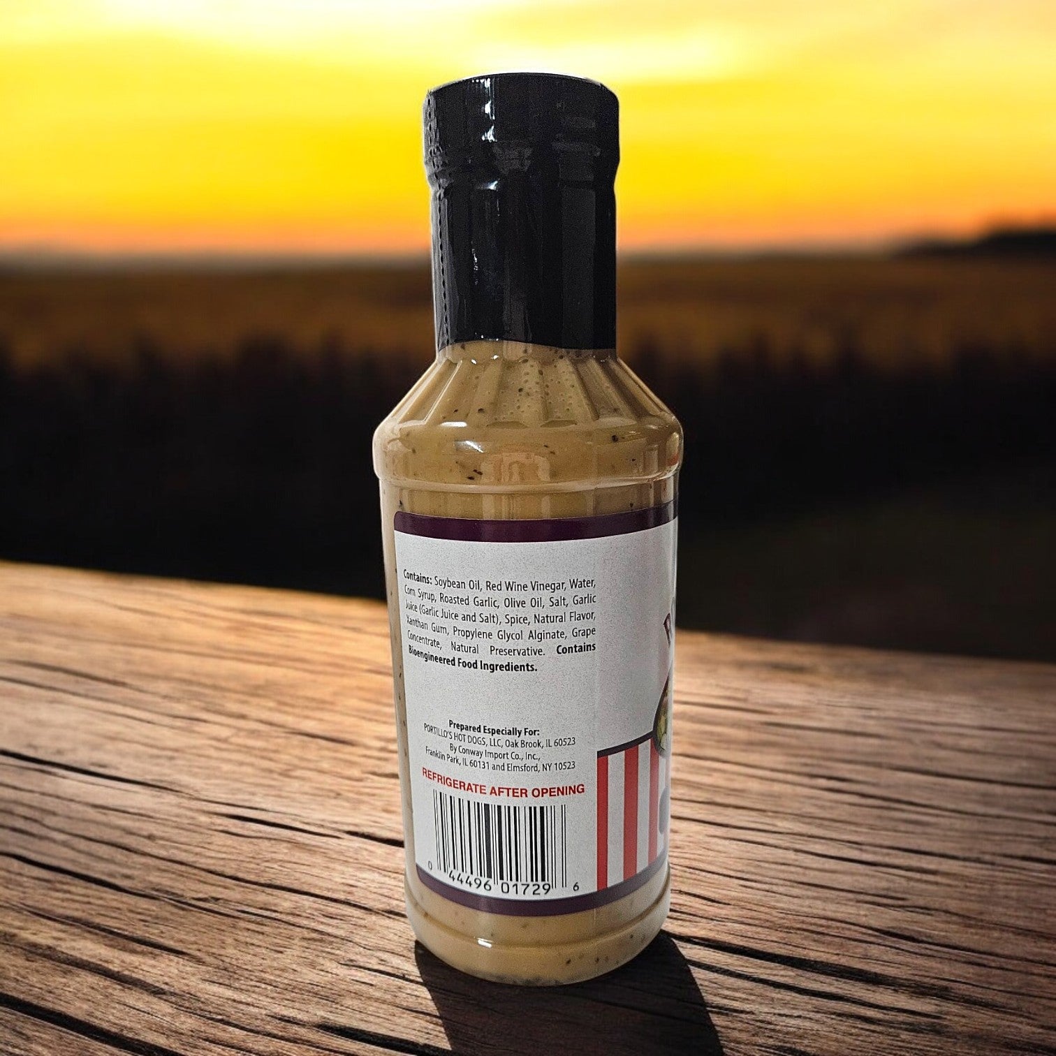 A 16 oz. Portillo's Roasted Garlic Vinaigrette bottle sits on a wooden surface, its label showcasing ingredients against a sunset backdrop.