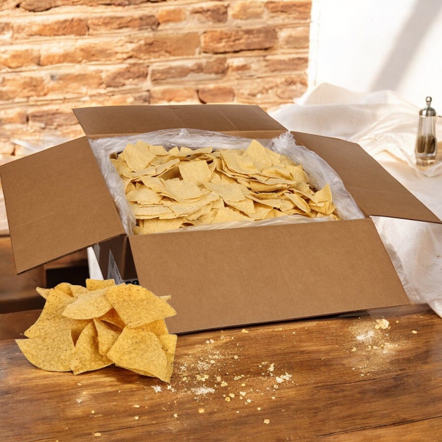 A cardboard box filled with El Milagro Corn Tortillas - 4-Way Precut Homestyle sits on a wooden table, with some chips scattered around. The rustic brick wall and nearby salt shaker evoke the authentic flavor of home-cooked meals.